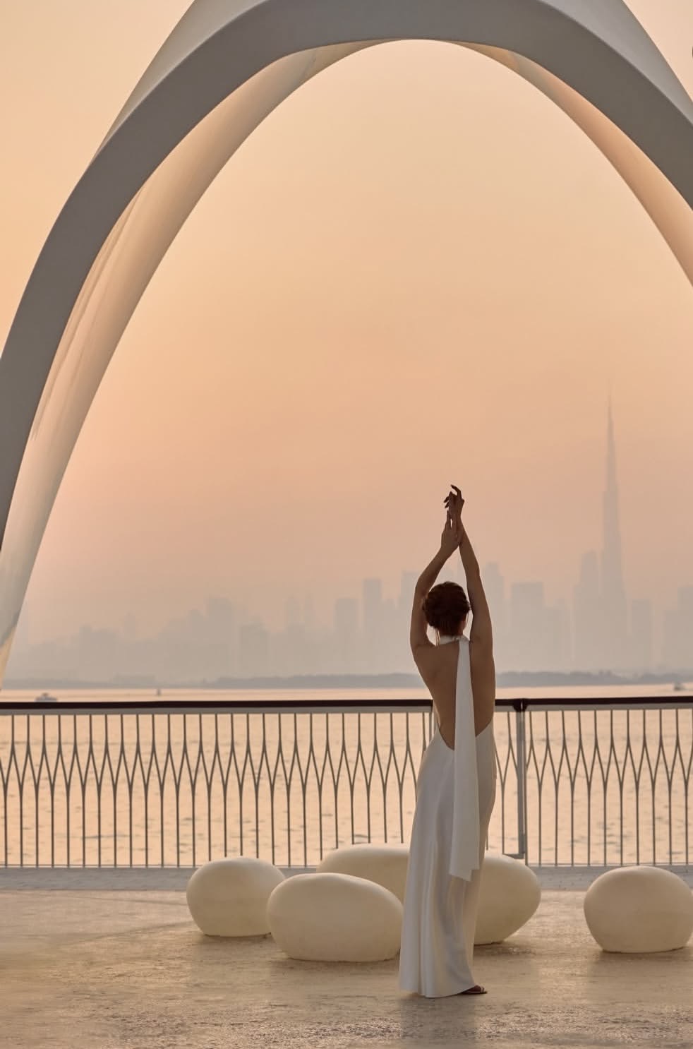 A woman in a white dress raising her arms under a white arch at Dubai Creek Harbour, the Burj Khalifa in the distance at sunset