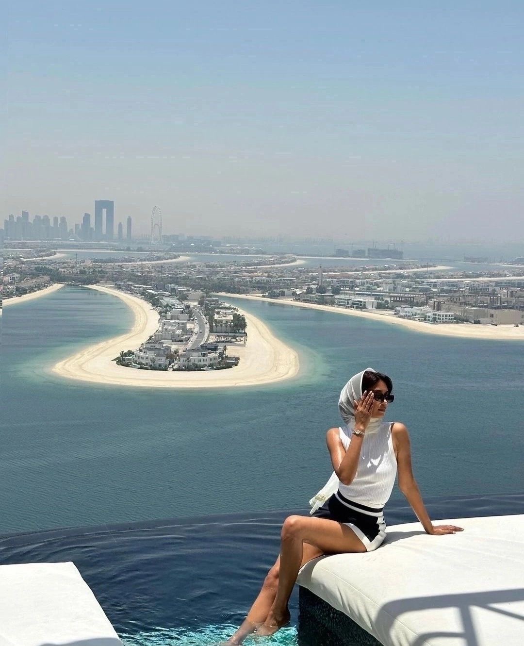 A woman in a white headscarf seated by an infinity pool overlooking Palm Jumeirah